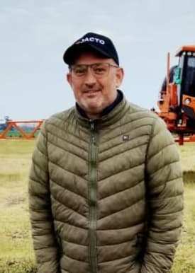 Roger, de Apa la Papa, en el campo con gorra y campera verde, con la pulverizadora detrás